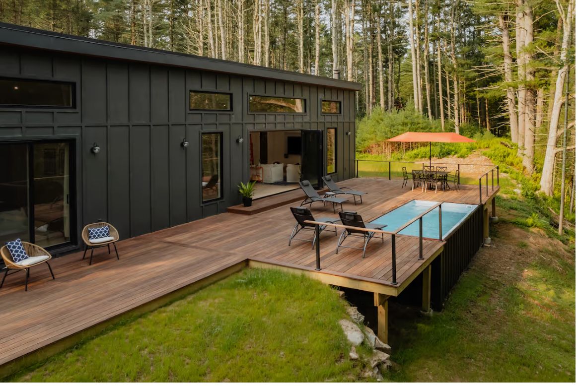 White Pines, a single-story black cabin with a wraparound deck and pool in a pine forest